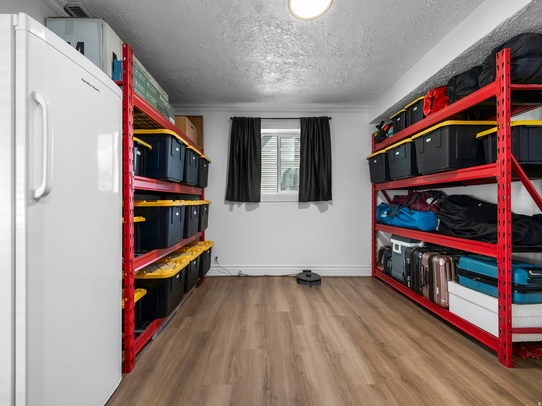 Utility room featuring wood-finish flooring, two rows of red-framed metal shelving, a single window with dark drapes, and a ceiling-mounted light fixture