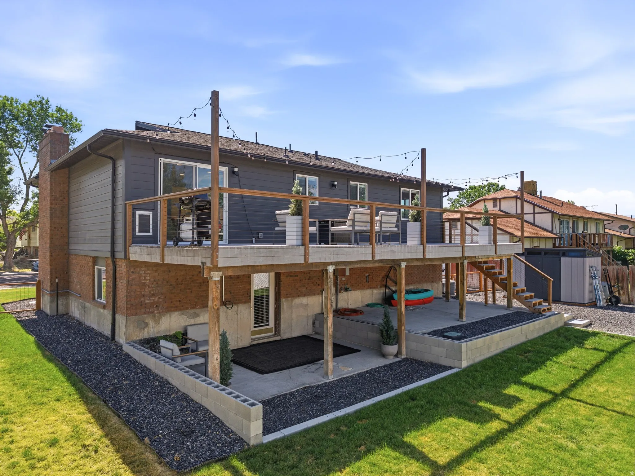 Rear exterior featuring a spacious elevated deck with wood railings and string lighting, a lower concrete patio, and a brick and siding facade