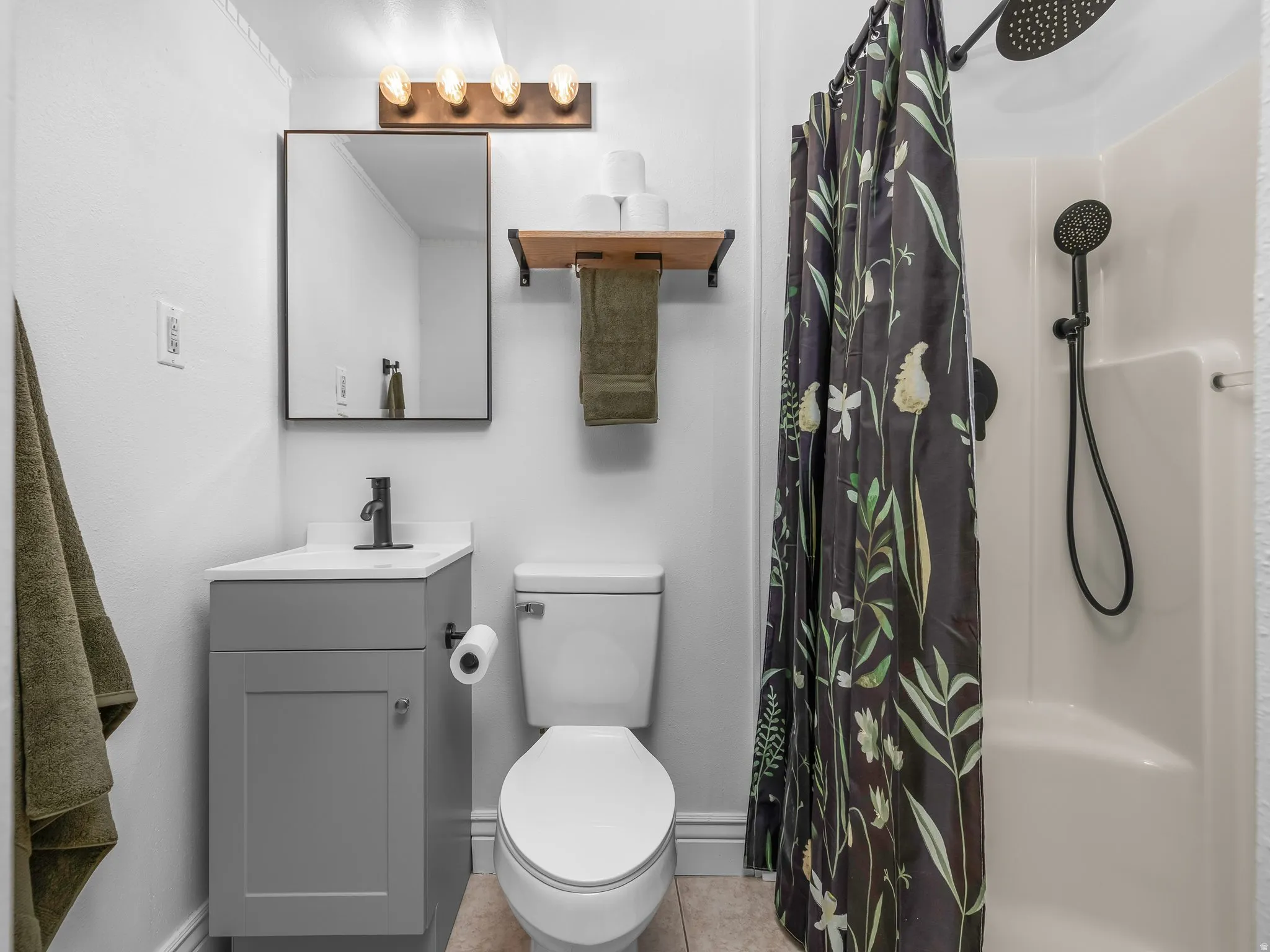 Bathroom featuring a single-piece shower surround with a dual-head matte black shower system, a gray vanity with a white countertop and matte black faucet, and a wall-mounted rectangular mirror with a four-bulb vanity light fixture