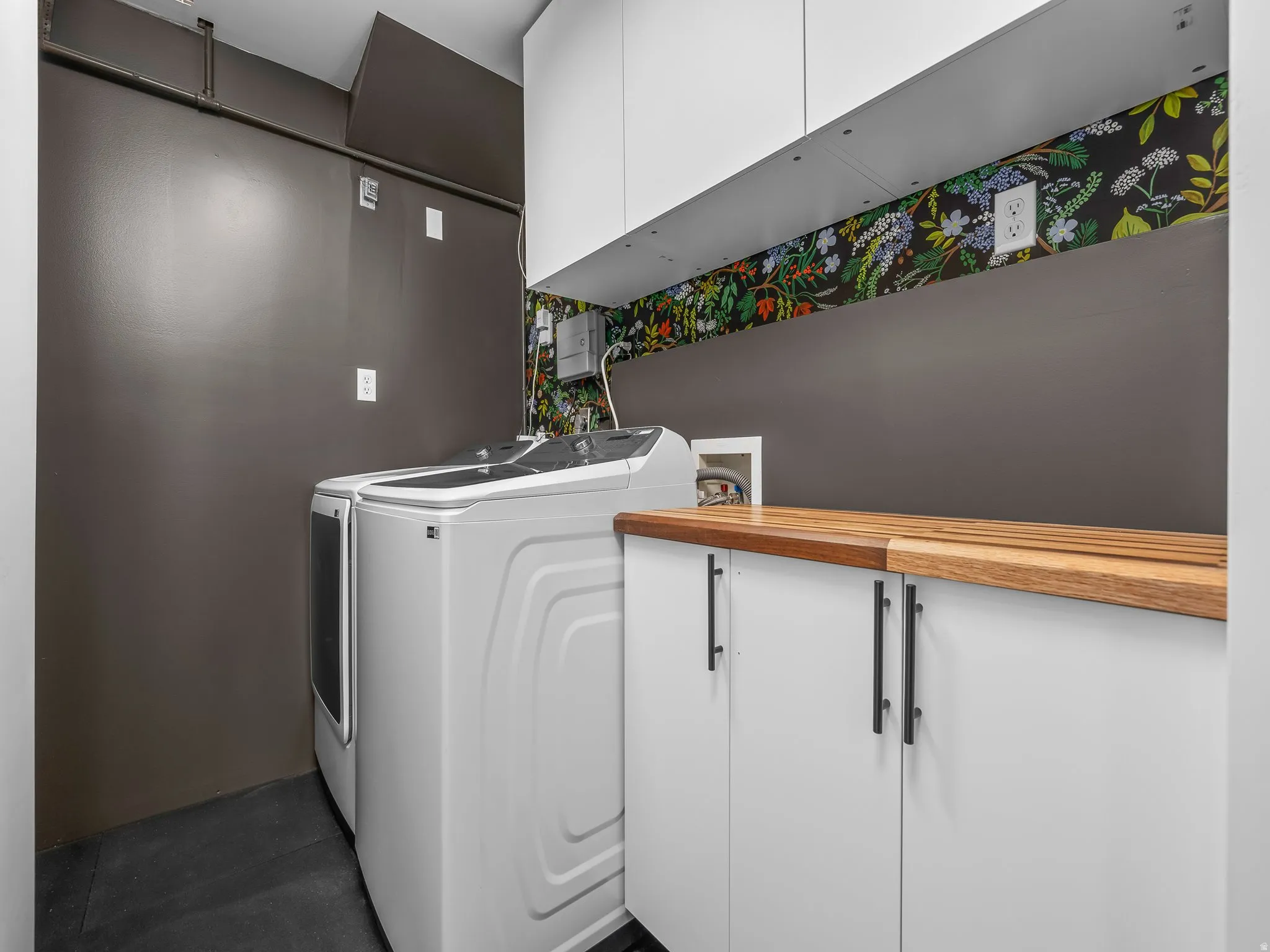 Dedicated laundry space featuring a wood-finish countertop, white cabinetry with black hardware, white overhead cabinets, and dark flooring