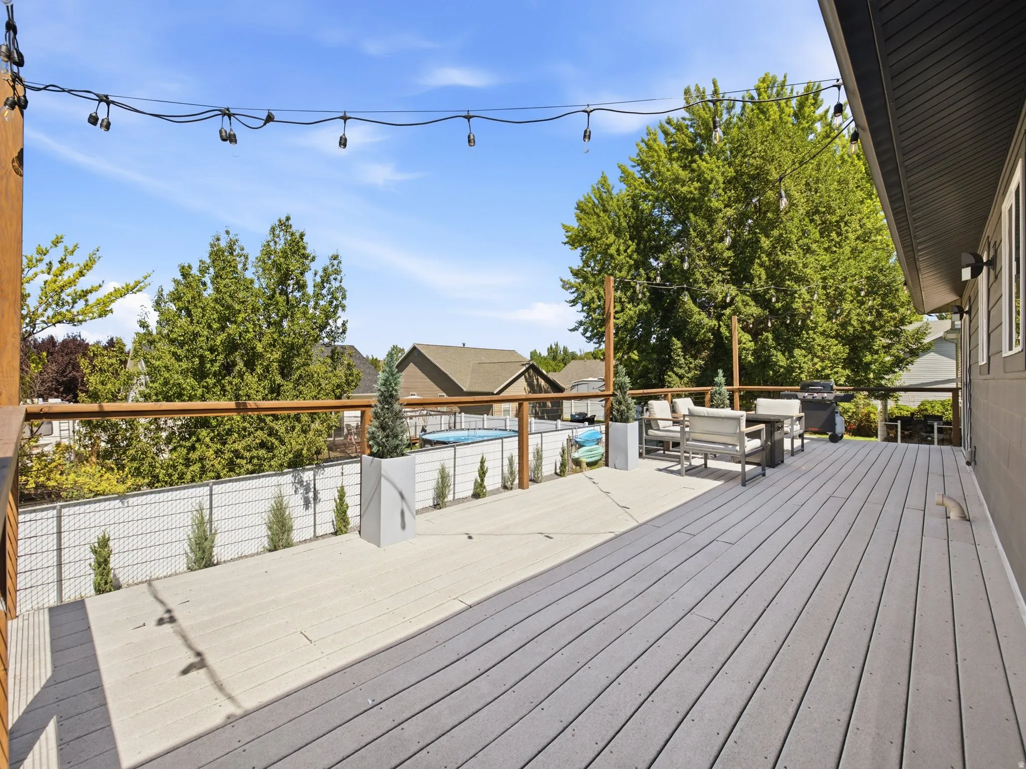 Spacious outdoor deck featuring two-tone composite decking, wood railing, string lighting, and a perimeter fence with mature trees