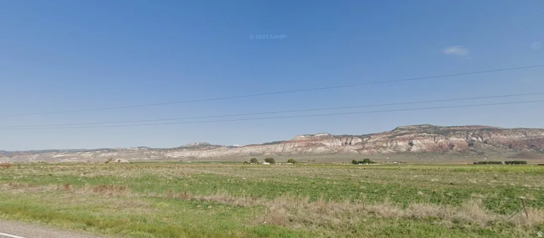 Expansive green fields under a clear blue sky, featuring distant mountains with visible rock formations and scattered structures in the midground