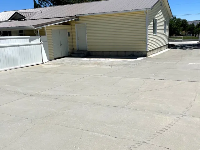 Spacious concrete driveway featuring a durable metal roof structure, light yellow siding, a white privacy fence, and a convenient exterior access door