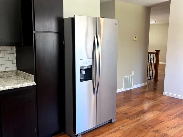 Stainless steel side-by-side refrigerator, dark wood-finish cabinetry, light-toned countertop, white subway tile backsplash, and wood-finish flooring