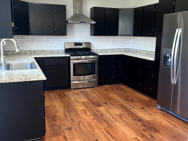 Modern kitchen featuring dark cabinetry, light speckled countertops, stainless steel appliances, a subway tile backsplash, and wood-finish flooring