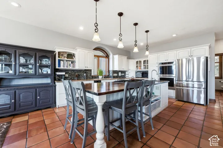 Kitchen featuring terracotta-style tile flooring, white cabinetry, stainless steel appliances, a center island with a wood-finish countertop, and pendant lighting