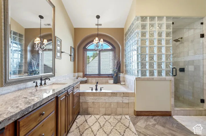 Bathroom featuring a dual vanity with a stone countertop and wood cabinetry, a built-in soaking tub with a tiled surround, an arched window with blinds, a glass block privacy wall, and a separate glass-enclosed shower