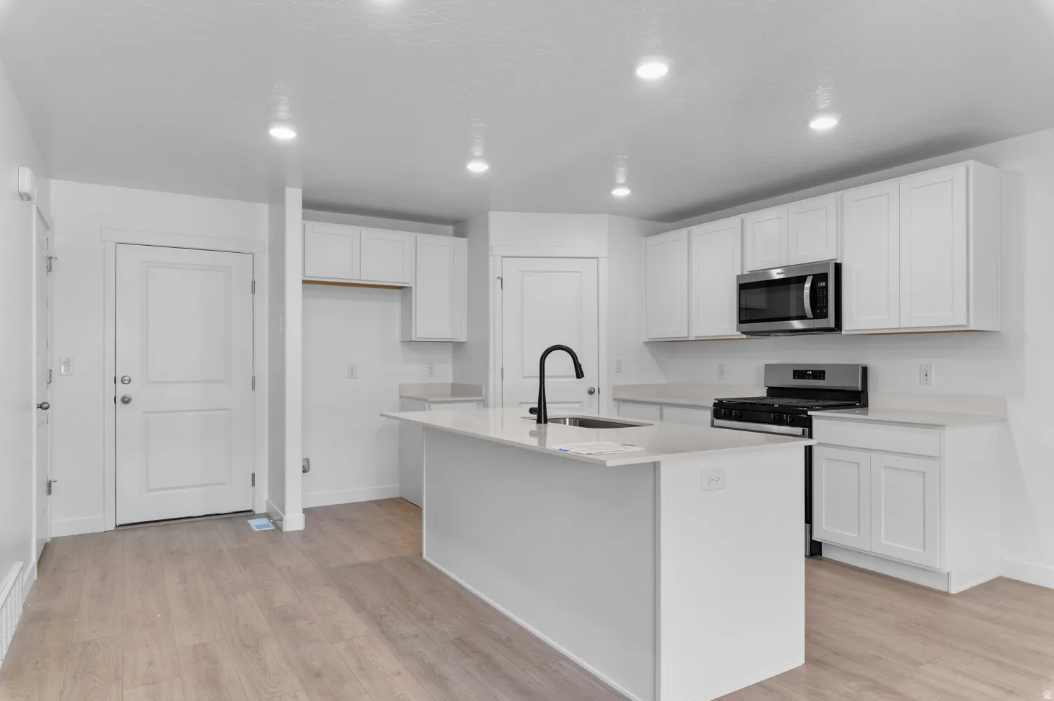 Modern kitchen featuring an island with an undermount sink and matte black faucet, white shaker-style cabinetry, stainless steel microwave, gas range, and light wood-finish flooring