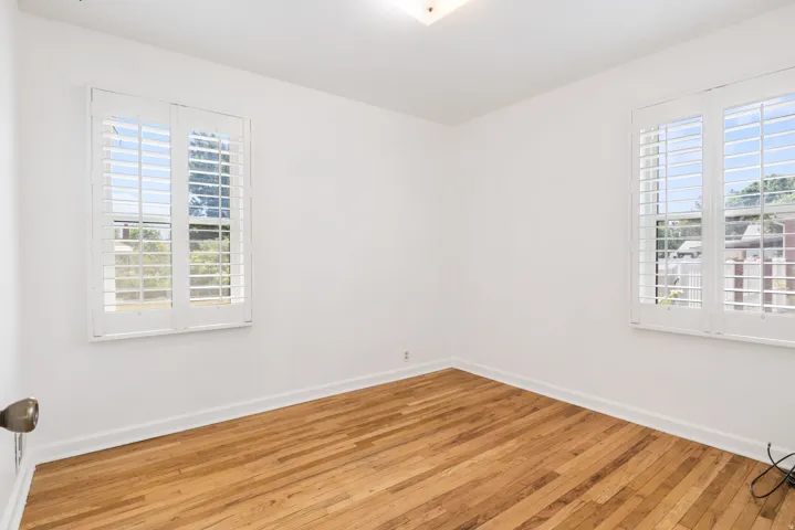 Bright room featuring two windows with white plantation shutters, wood-finish flooring, white walls, and a ceiling-mounted light fixture