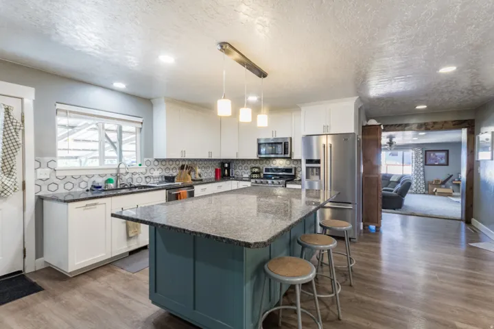 Open-concept kitchen featuring an oversized island with a stone-finish countertop, white cabinetry with contemporary hardware, stainless steel appliances, and wood-finish flooring