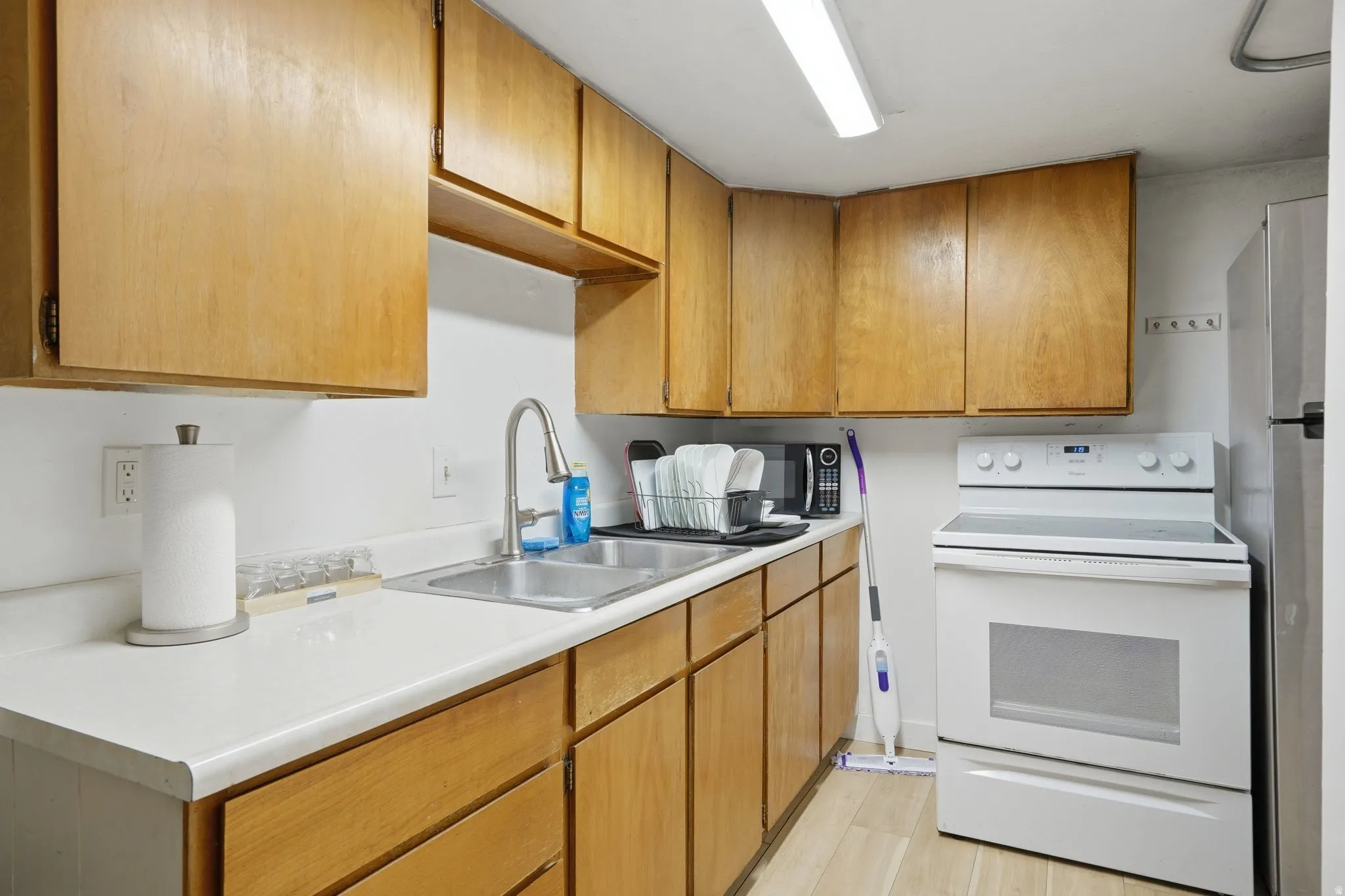 Kitchen featuring wood cabinetry, light-toned countertops, a stainless steel double basin sink with a high-arch faucet, a white electric range, and light wood-finish flooring