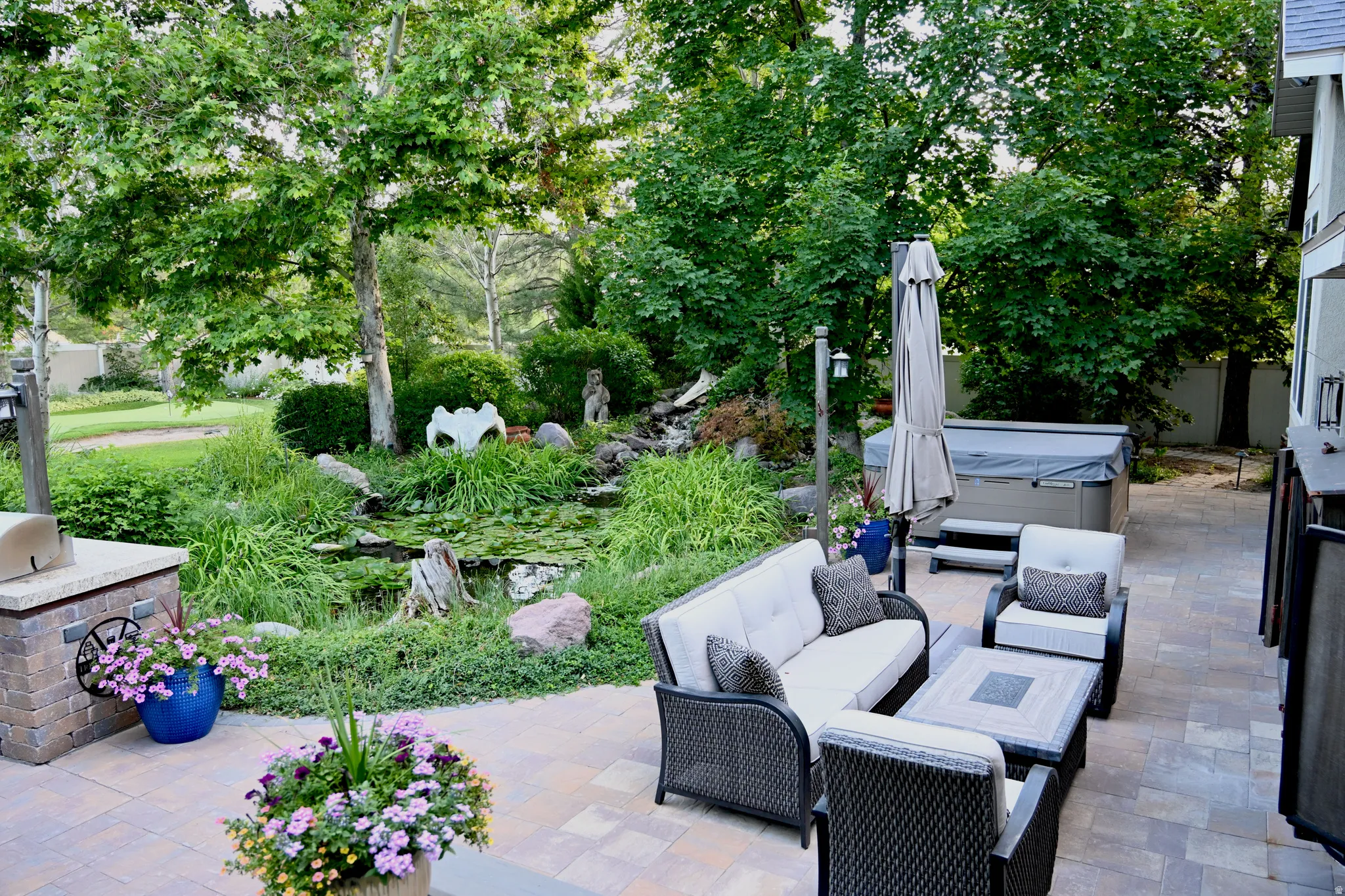 Paver patio with an outdoor kitchen, covered spa, and lush landscaping featuring a pond