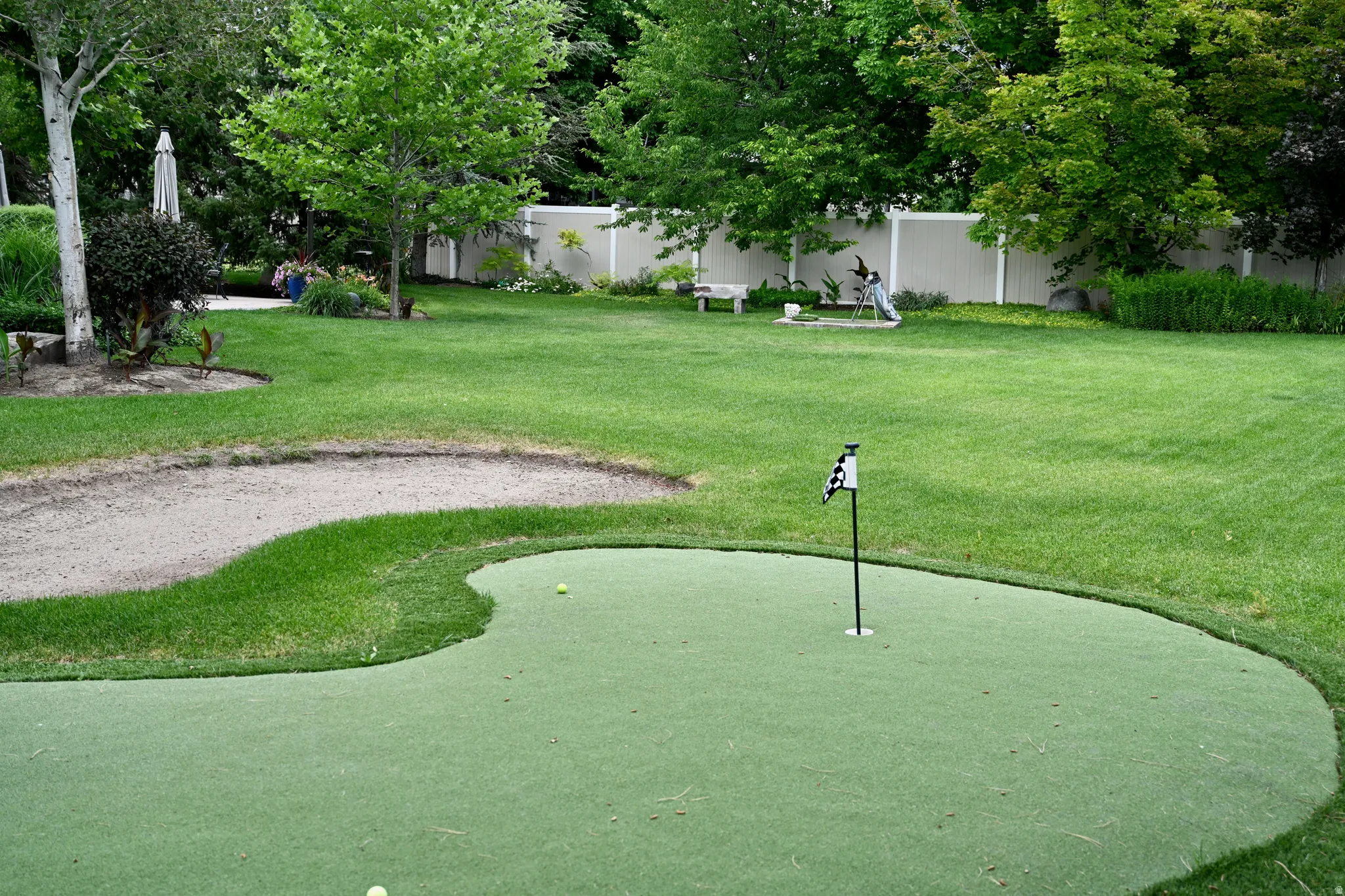 Expansive lawn featuring a putting green with flag, a sand trap, mature trees, and a privacy fence