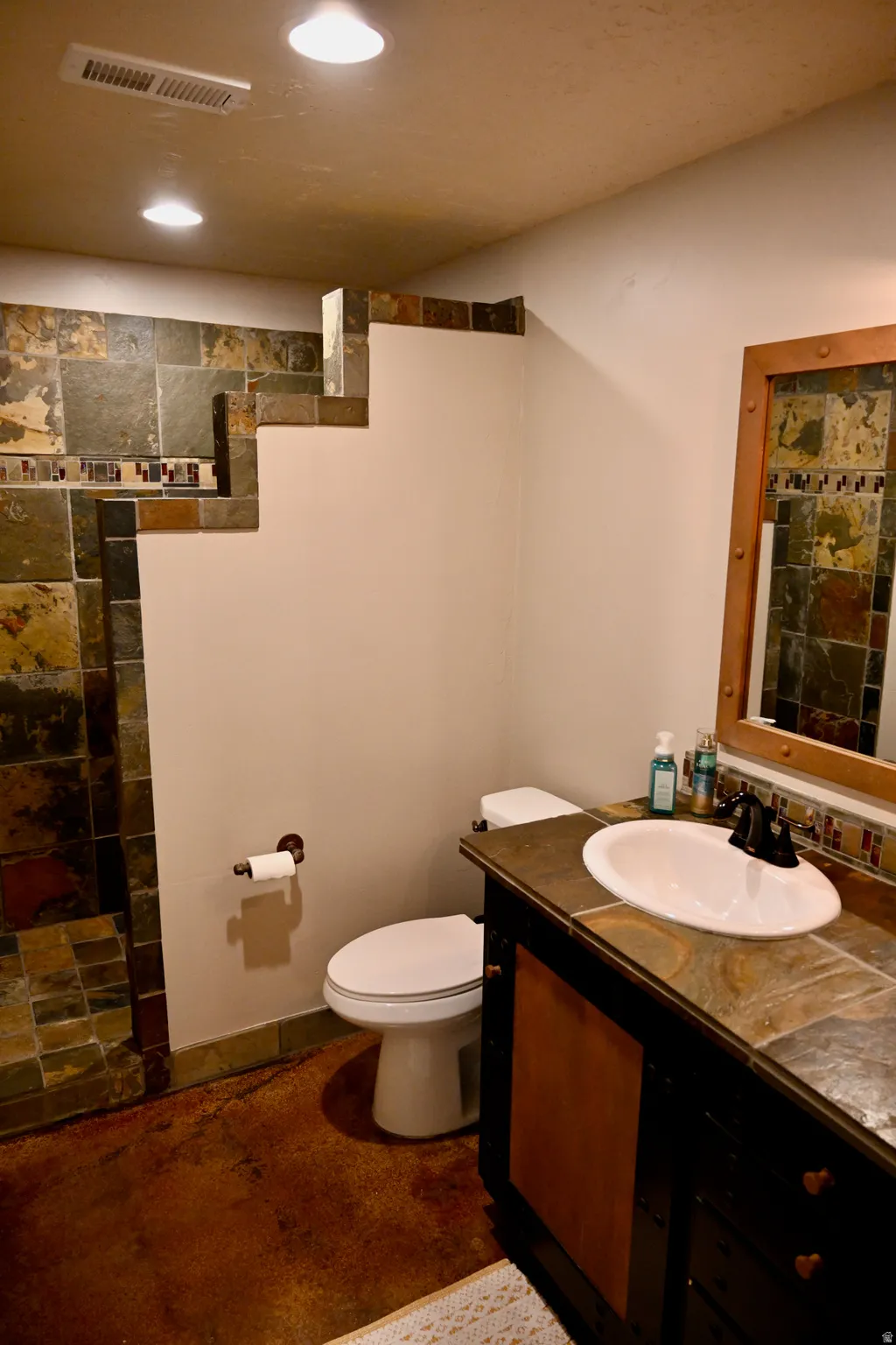 Bathroom featuring a tile vanity with an integrated sink and dark cabinet, stone tile shower, and a white commode