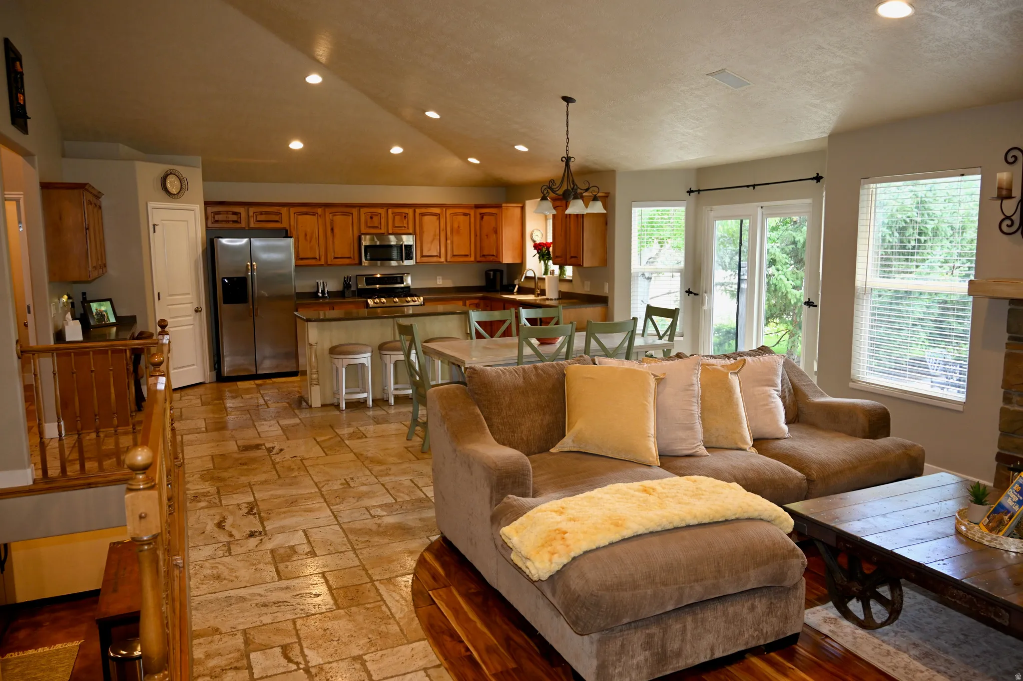 Expansive living area featuring vaulted ceilings with recessed lighting, large windows, a stone-clad fireplace, and a kitchen with wood cabinetry and stainless steel appliances