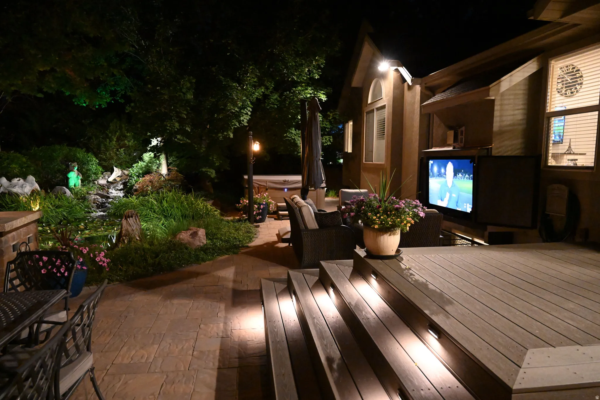 Expansive outdoor living area featuring a multi-level deck with integrated stair lighting, a stone-finish patio, a built-in outdoor television, and a lush landscaped garden with a water feature