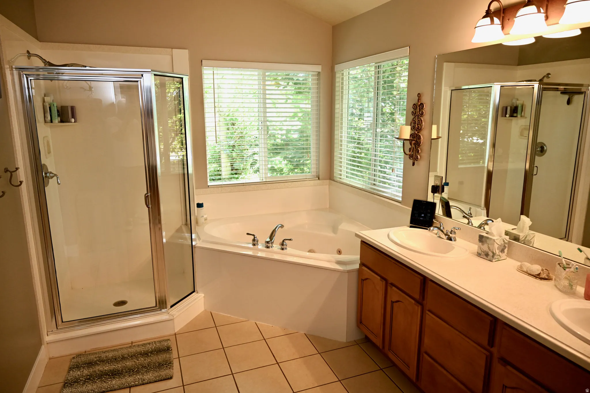 Bathroom featuring a corner soaking tub, dual windows with blinds, a separate glass-enclosed shower, a double vanity with integrated sinks, and neutral tile flooring