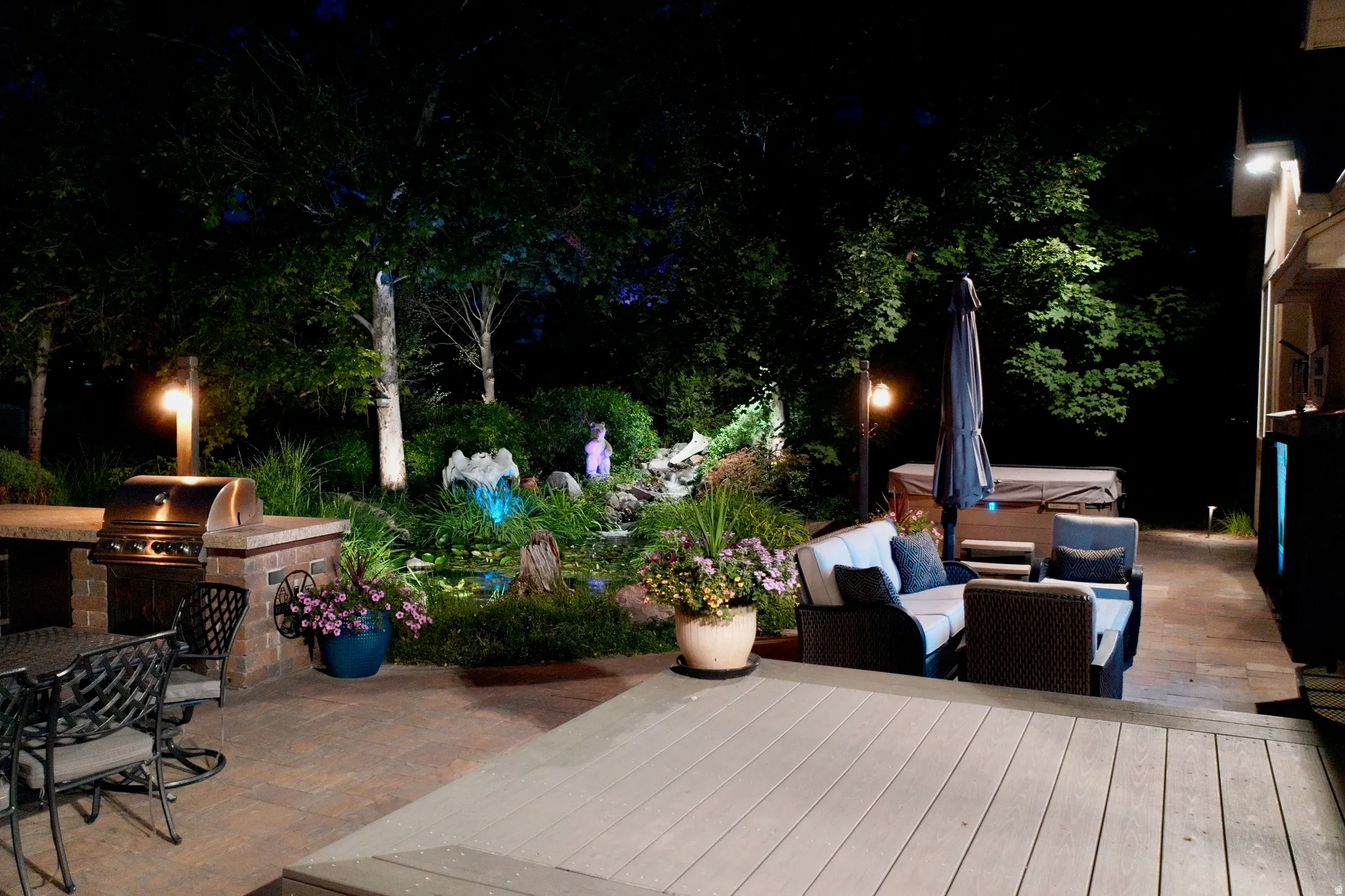Expansive outdoor living area featuring a built-in grill station, paver patio, composite decking, and a water feature with accent lighting