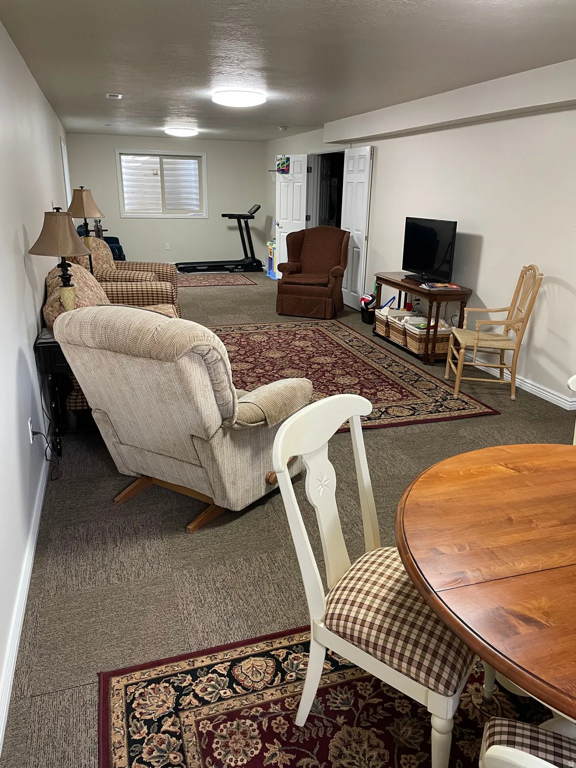 Carpeted recreation room featuring overhead flush-mount lighting, a window with horizontal blinds, and white bi-fold doors