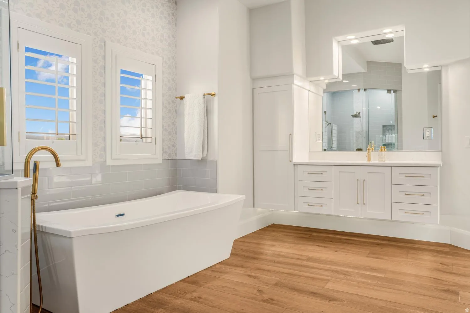 Spacious bathroom featuring a freestanding soaking tub with a floor-mounted golden faucet, dual shuttered windows, and wood-finish flooring