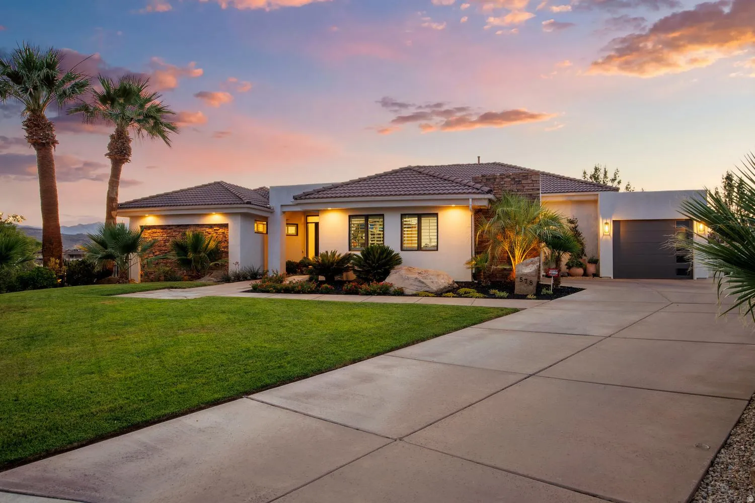 Contemporary stucco exterior featuring a tile roof, a two-car garage with a modern door, a manicured front lawn, and mature palm trees