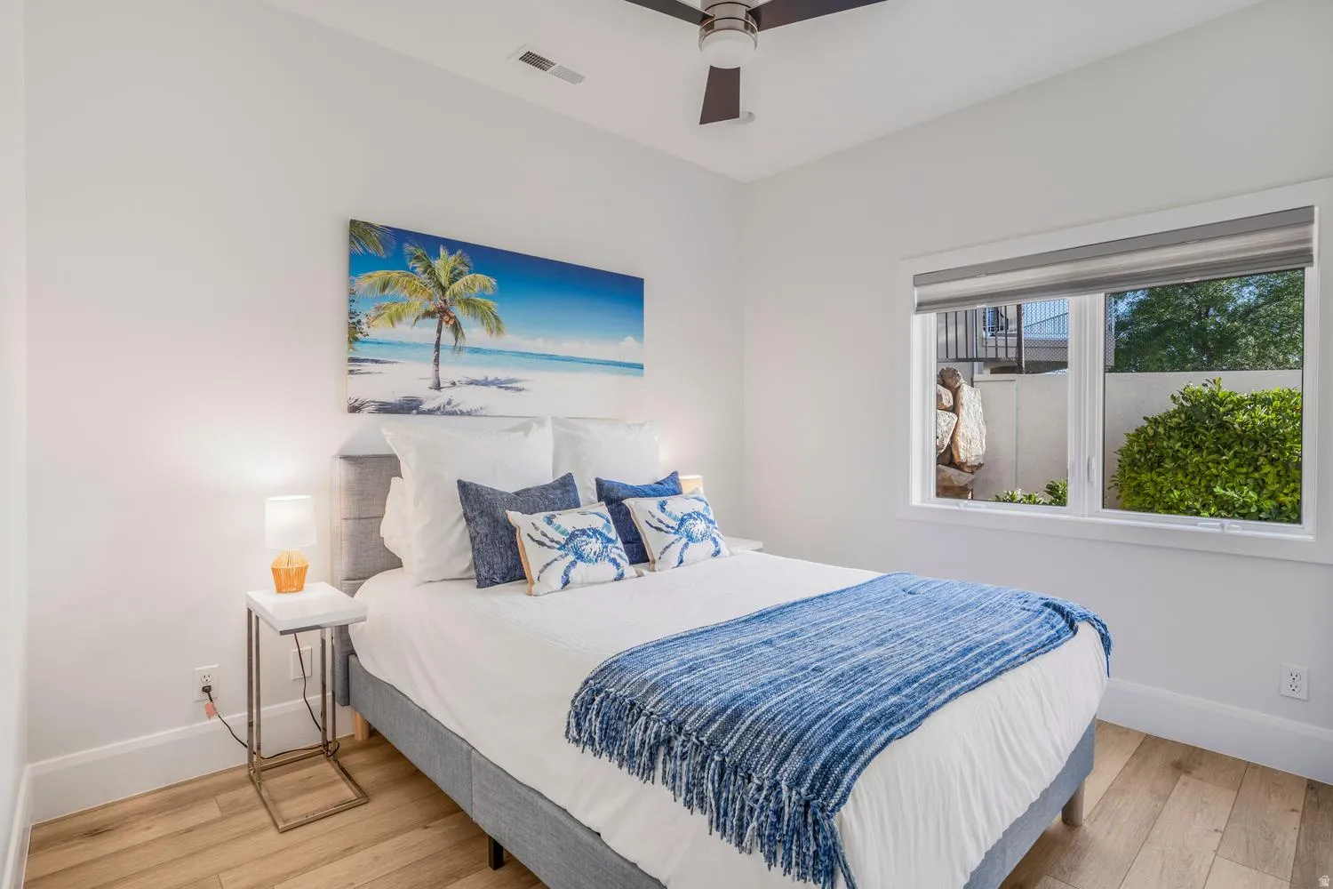 Bright bedroom featuring light wood-finish flooring, white walls, a ceiling fan, and a large window with roller shades
