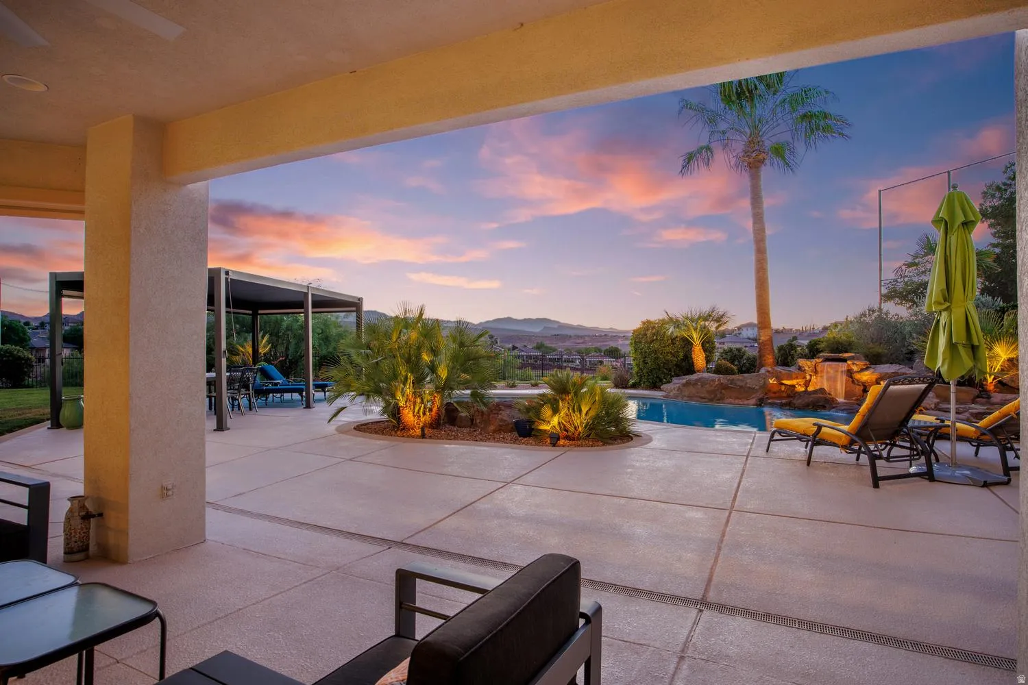 Expansive covered patio featuring a concrete surface, swimming pool with a rock waterfall, and mature palm trees