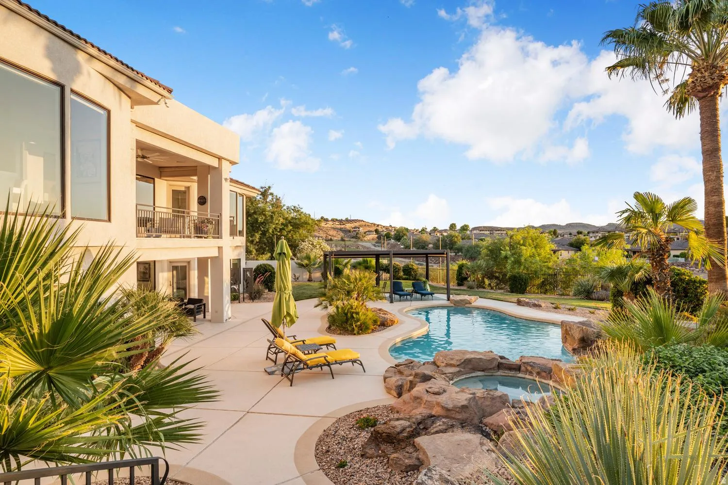 Expansive backyard featuring a custom-shaped swimming pool with a rock-surrounded spa, extensive concrete patio, mature palm trees, and established landscaping