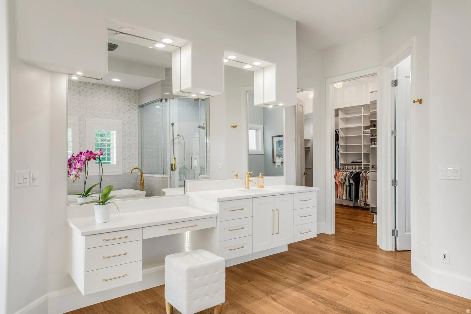 Bathroom featuring dual vanities with white cabinetry and gold-finish hardware, a built-in vanity desk, and wood-finish flooring