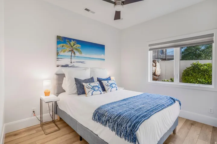 Bright bedroom featuring light wood-finish flooring, white walls, a ceiling fan, and a large window with roller shades