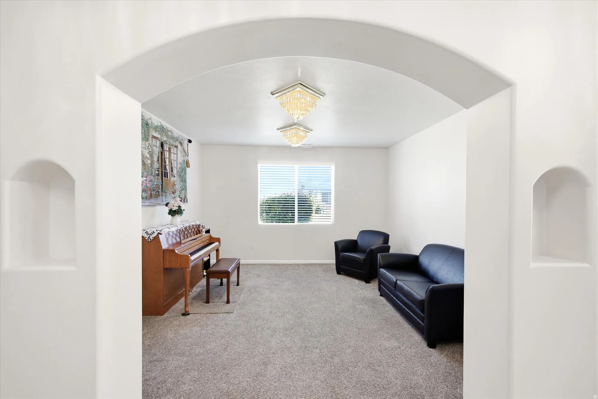 Archway entry with integrated display niches opens to a versatile room featuring two flush-mount crystal chandeliers, a large window with blinds, and neutral carpeting