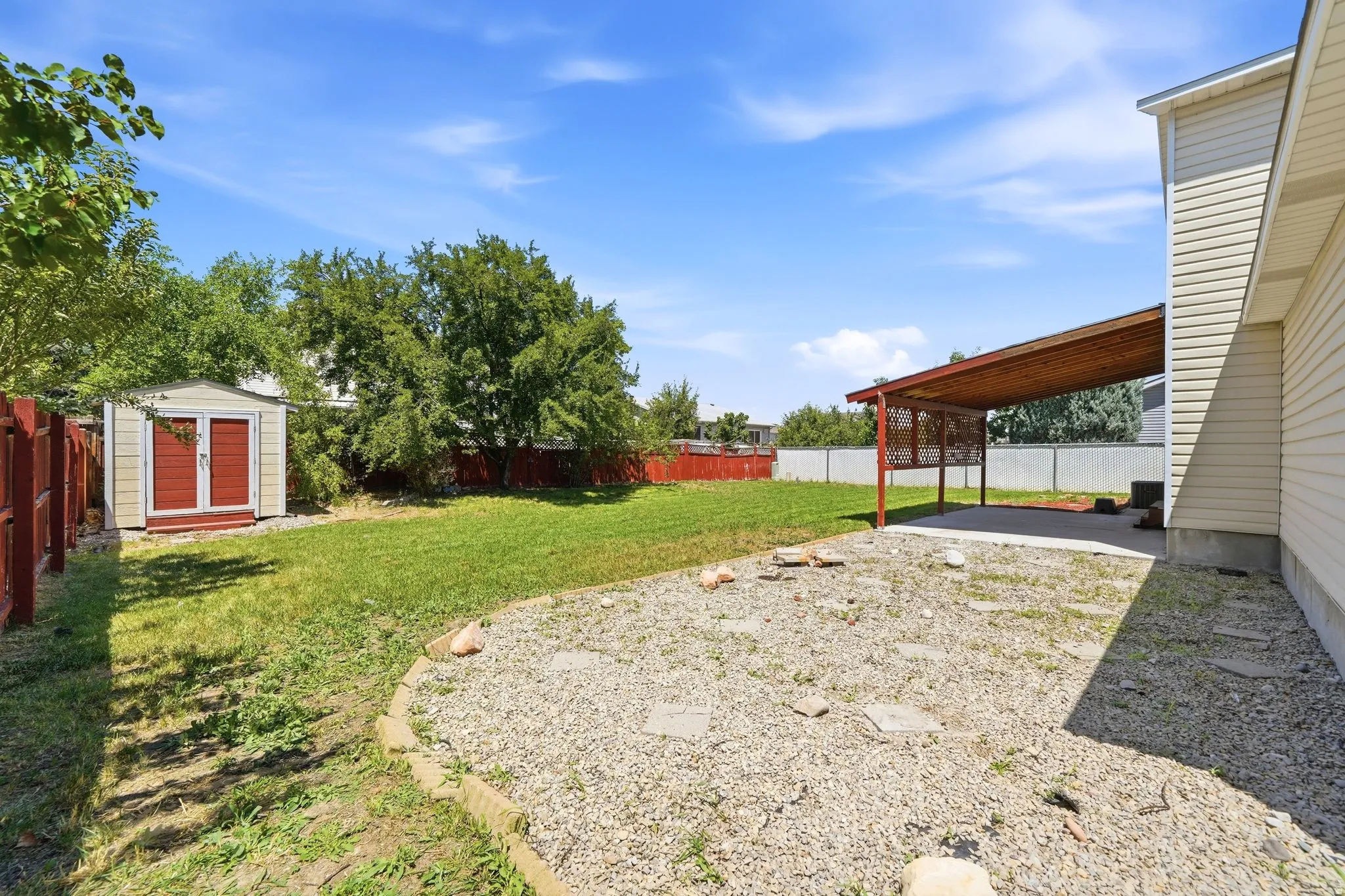 Expansive backyard featuring a covered patio with wood pergola, gravel landscaping, a grass lawn, and a storage shed with red doors
