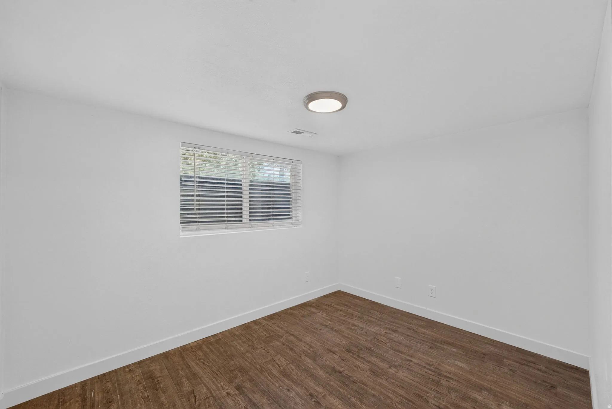 Room featuring neutral white walls, wood-finish flooring, a window with horizontal blinds, and a flush-mount ceiling light fixture