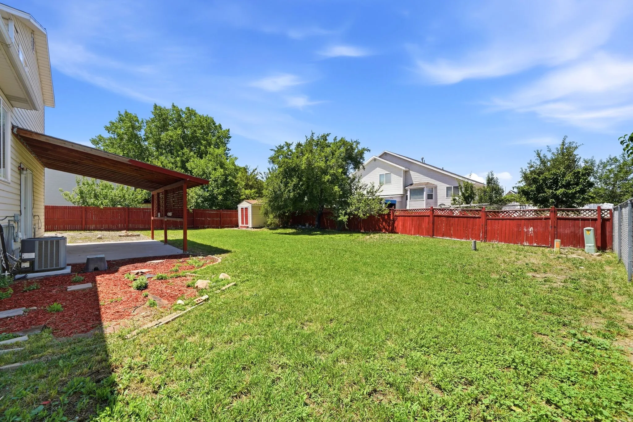 Expansive green yard with a covered patio, a red wooden privacy fence, and a storage shed