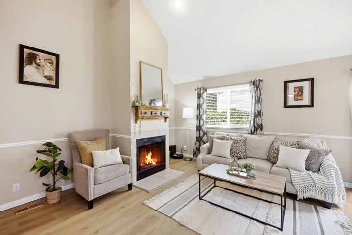 Living area featuring vaulted ceilings, light wood-finish flooring, a stone fireplace with a decorative mantel, and a large window providing natural light