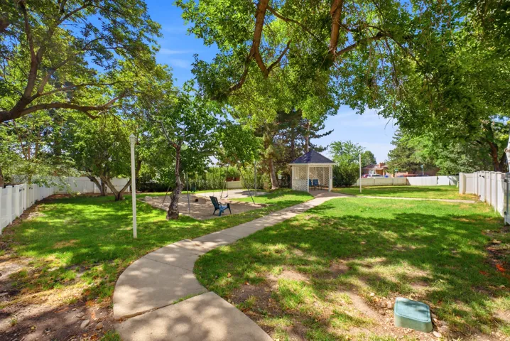 Expansive green space featuring a white gazebo with a dark roof, mature trees providing natural shade, a meandering concrete pathway, and white privacy fencing