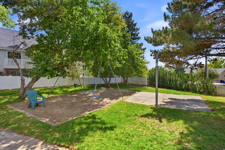 Expansive outdoor area featuring a swing set over a sand base, mature trees providing shade, a concrete pad, and a white privacy fence