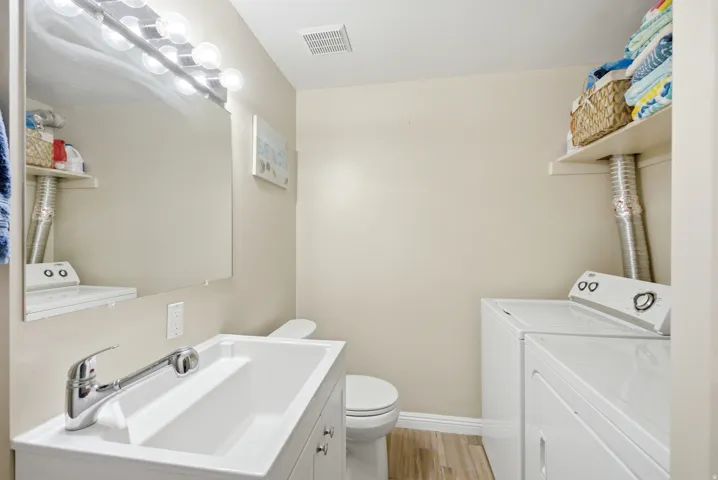 Bathroom featuring a vanity with an integrated sink and chrome faucet, a wall-mounted mirror with overhead lighting, a toilet, and light wood-finish flooring