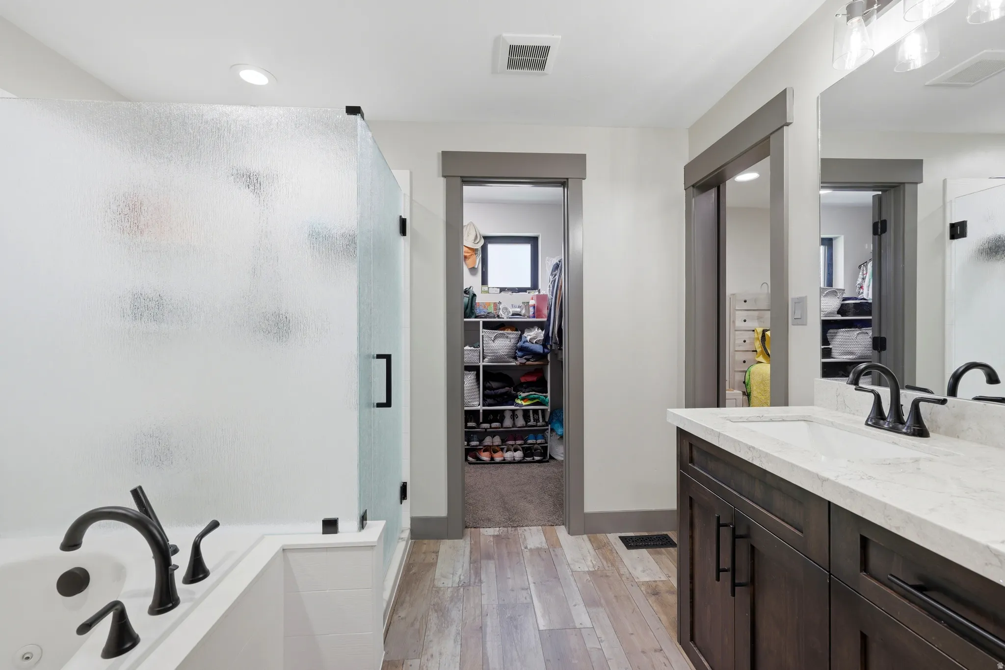 Full bathroom featuring a rectangular drop-in tub with matte black fixtures, a walk-in shower with frosted glass enclosure, a single vanity with a white countertop and undermount sink, and wood-finish flooring