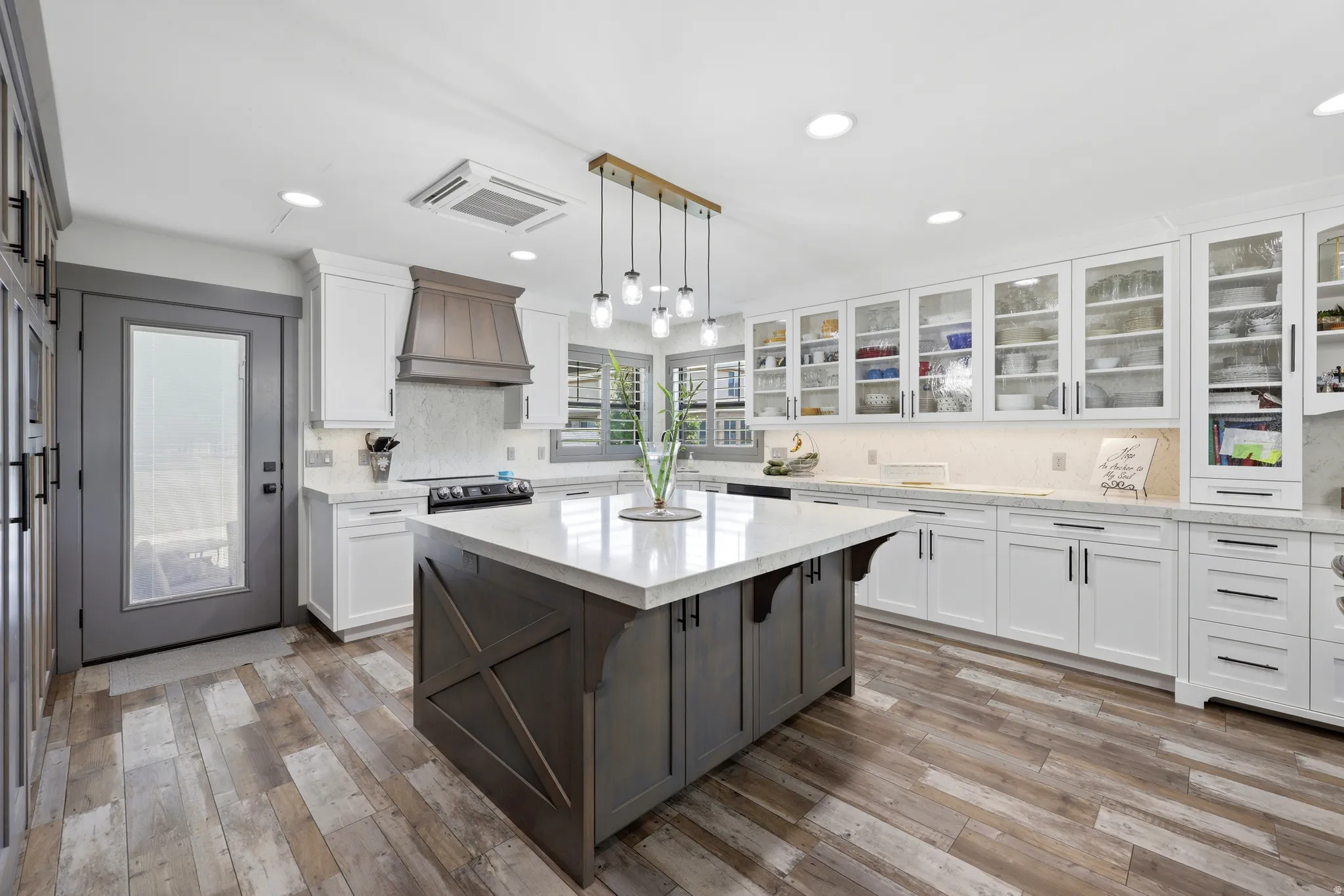 Spacious kitchen featuring a large central island with a light-toned countertop, wood-finish flooring, white cabinetry with dark hardware, a wood-paneled range hood, and extensive upper glass-front cabinets