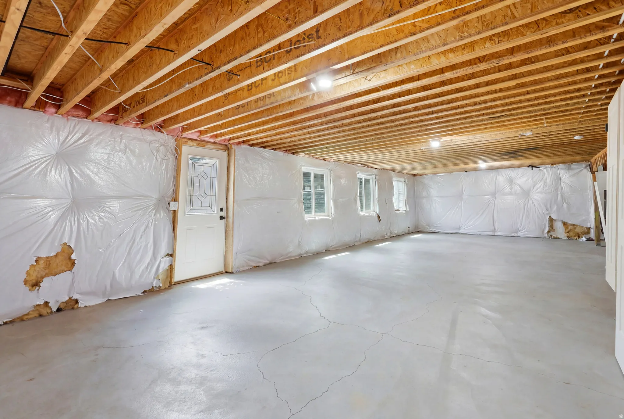 Expansive unfinished basement featuring a concrete floor, exposed wood ceiling joists, three windows, recessed lighting, and a white exterior door with decorative glass