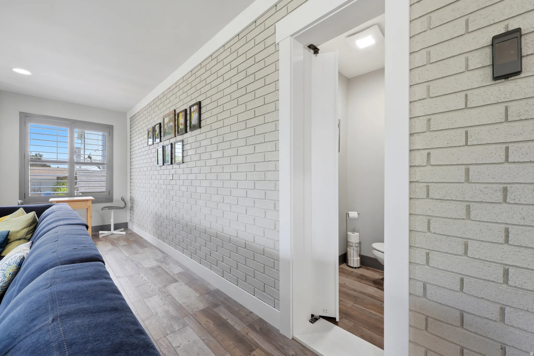 Interior space featuring light gray brick-patterned accent wall, wood-finish flooring, white trim, and a window with plantation shutters