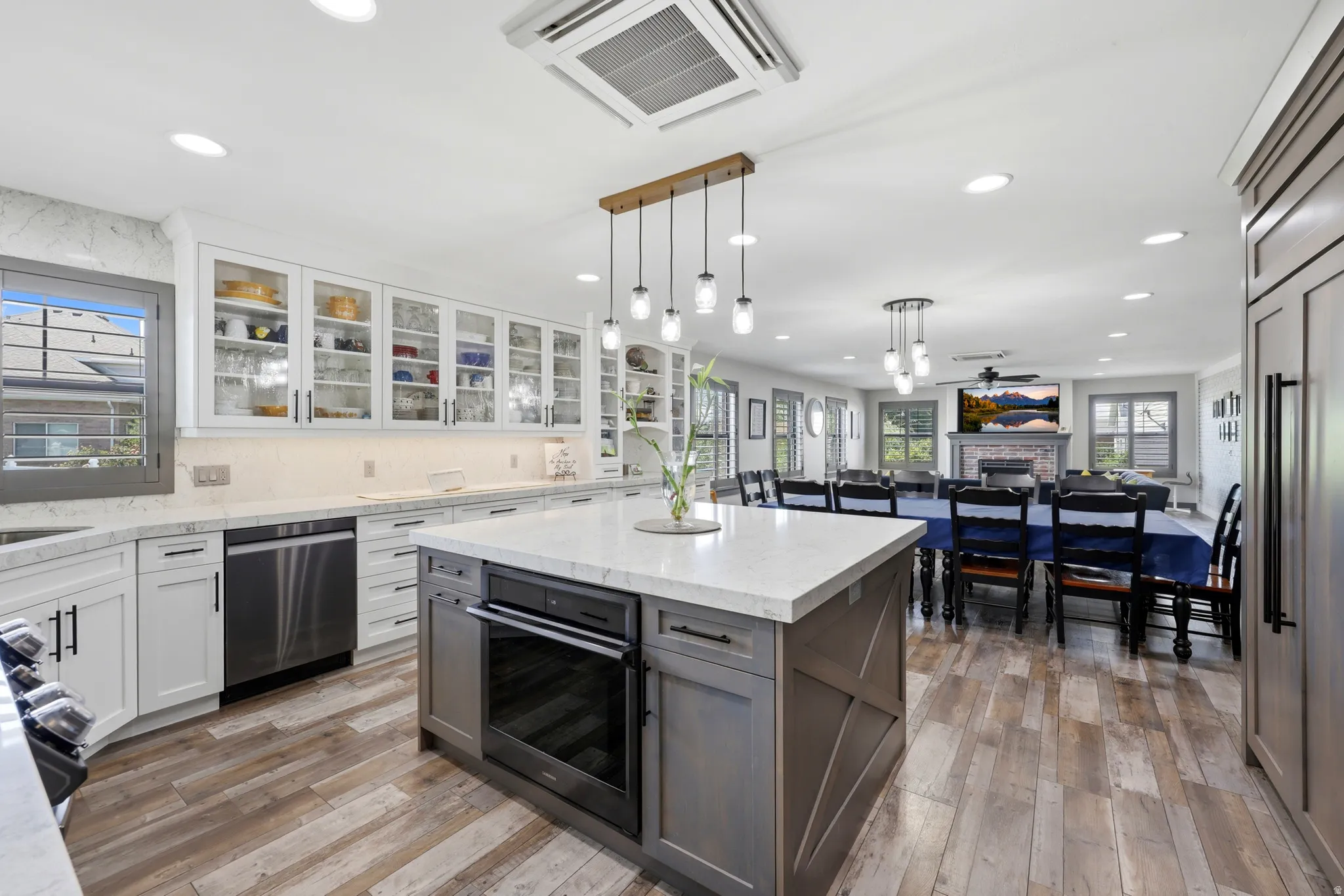 Expansive kitchen featuring a large central island with an integrated oven, wood-finish flooring, white cabinetry with glass-front uppers, stainless steel appliances, and recessed lighting