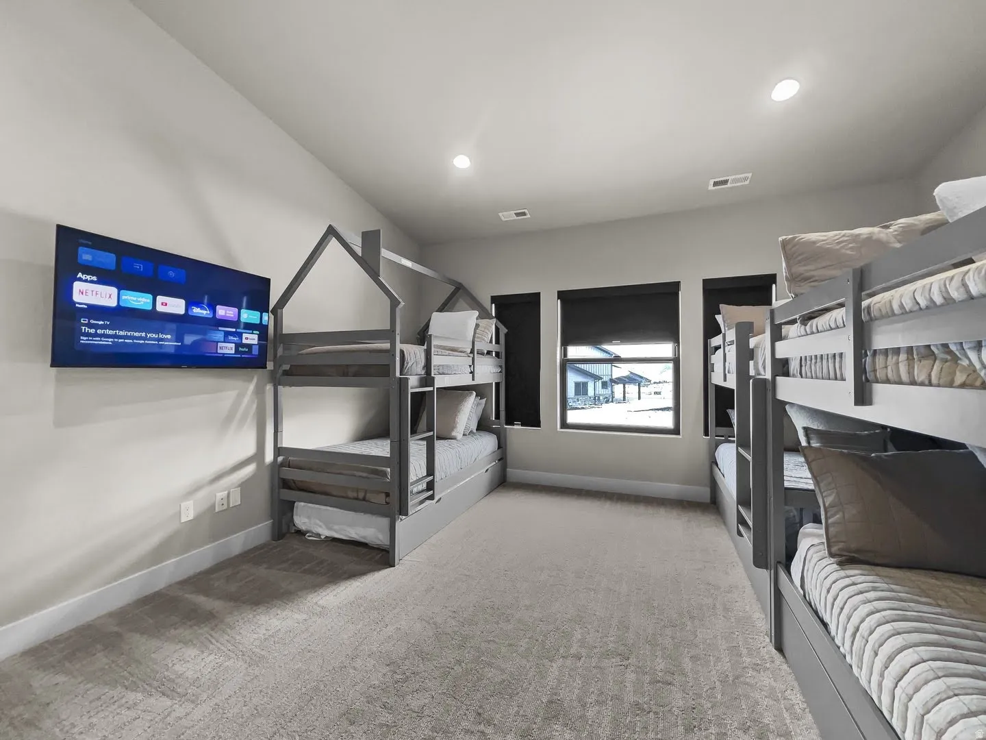 Room featuring built-in bunk beds with house-shaped frames, a wall-mounted television, multiple windows with dark roller shades, recessed lighting, and textured carpet flooring