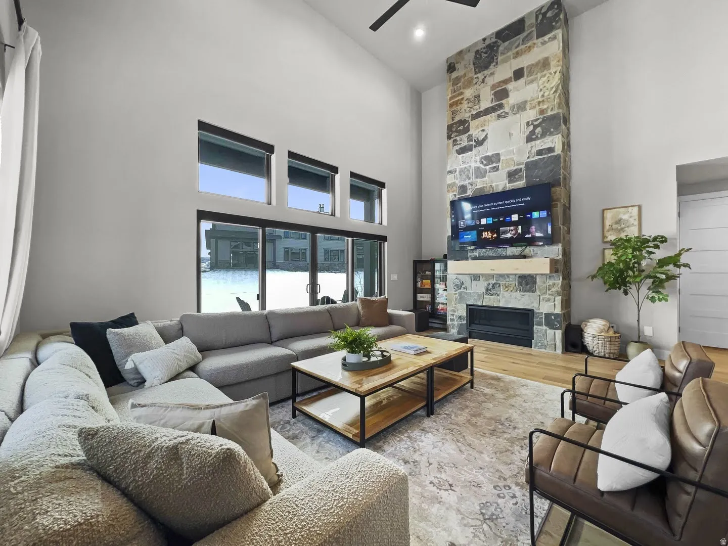 Vaulted ceiling living area featuring a floor-to-ceiling stone-faced fireplace with a wood mantle, a ceiling fan, wood-finish flooring, and expansive sliding glass doors with transom windows