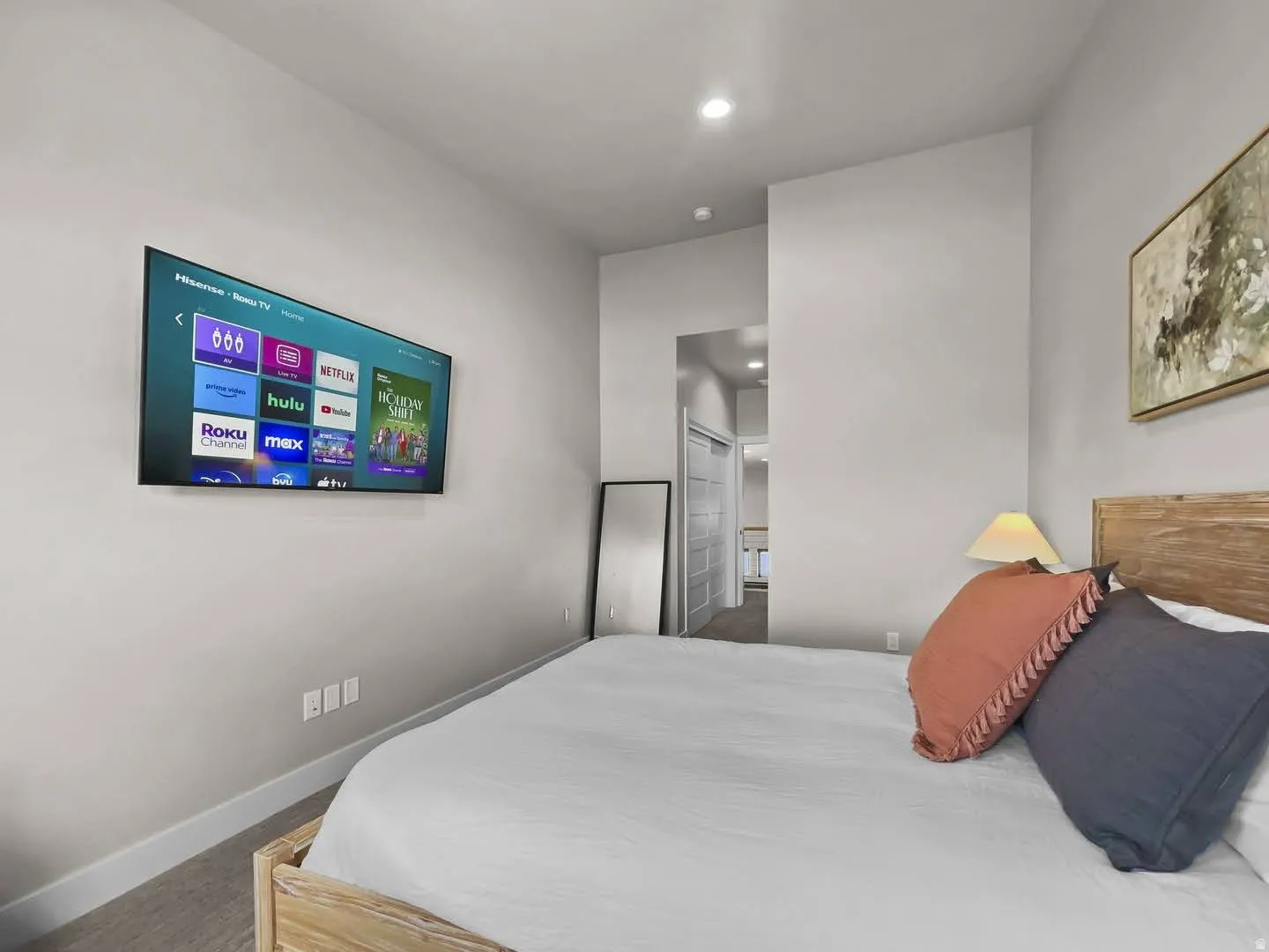 Neutral-toned room featuring a mounted flat-screen television, textured carpeting, and a light wood-finish headboard