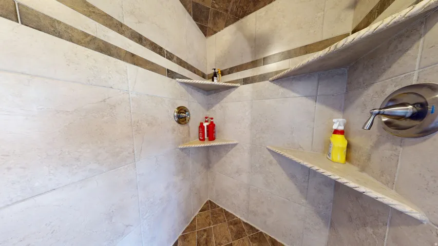 Tiled shower enclosure featuring light-toned large format wall tiles with horizontal accent bands, a dark-toned diagonal grid floor tile, multiple built-in corner shelves, and chrome fixtures