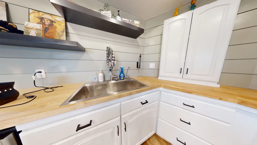 Corner kitchen sink with a stainless steel basin, wood-finish countertops, white shaker-style cabinetry with dark hardware, and horizontal plank wall paneling