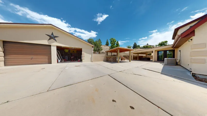 Expansive concrete patio area featuring a detached garage with two bays, an outdoor covered structure with a stone base, and a covered patio extending from the main residence
