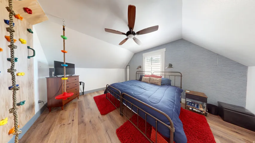 Attic room featuring wood-finish flooring, a sloped ceiling with a ceiling fan, a window with white shutters, and a climbing wall with colorful holds
